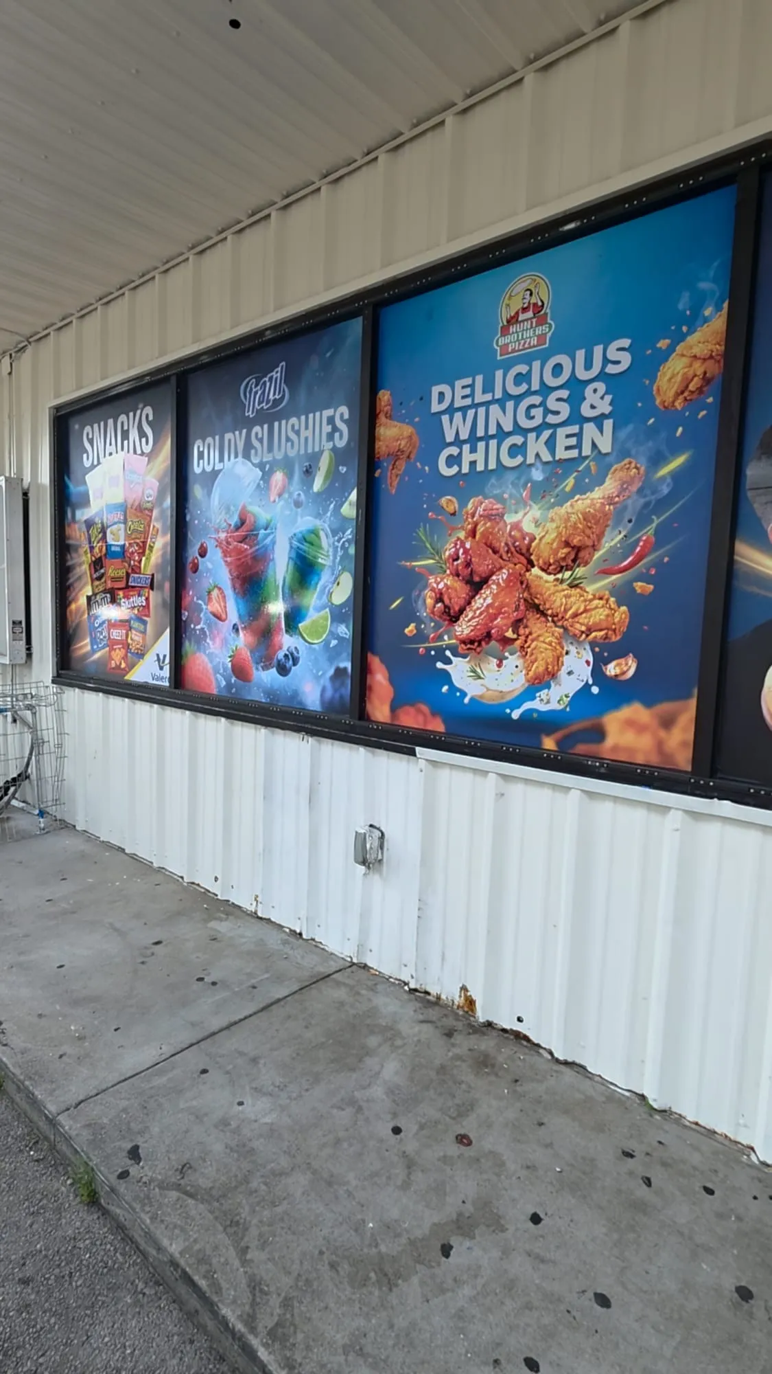 Row of large printed window panels on a white metal building showing snacks, colorful slushies and a large photo of fried chicken wings.