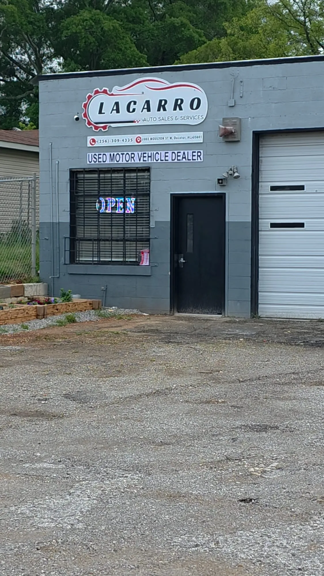 Gray block building with a white custom-shaped sign mounted high on the wall, a long strip sign below it, a barred window with a lit open sign, a dark door and a roll-up door.