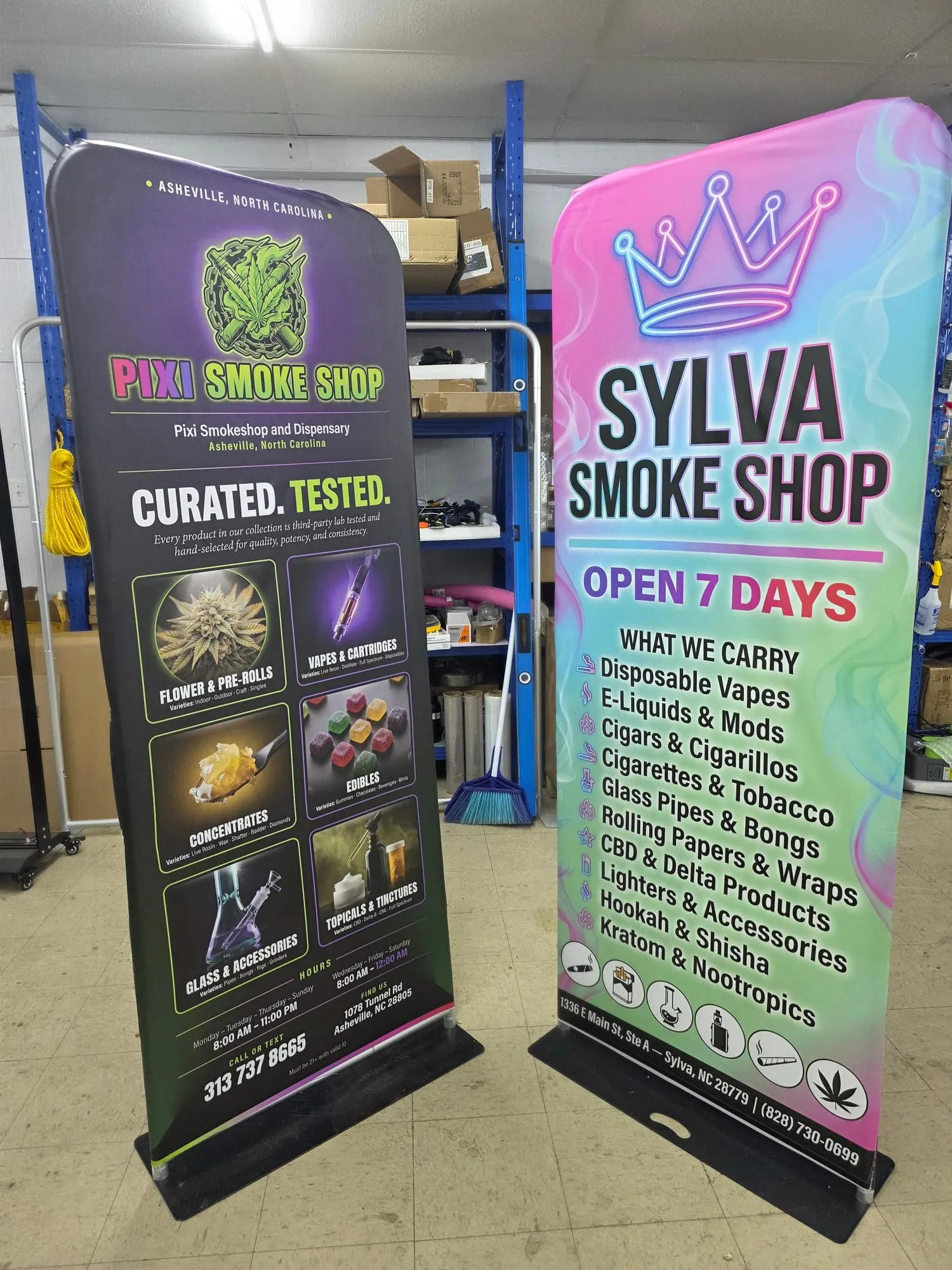 Two tall tension fabric stands side by side on a tile floor in a workshop, one dark purple with product tiles, one pink and green with a crown graphic and a list of items.