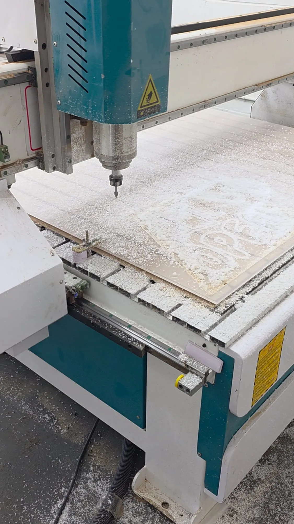 A CNC router with its bit above a panel on the cutting bed, the bed and the panel covered in white shavings.