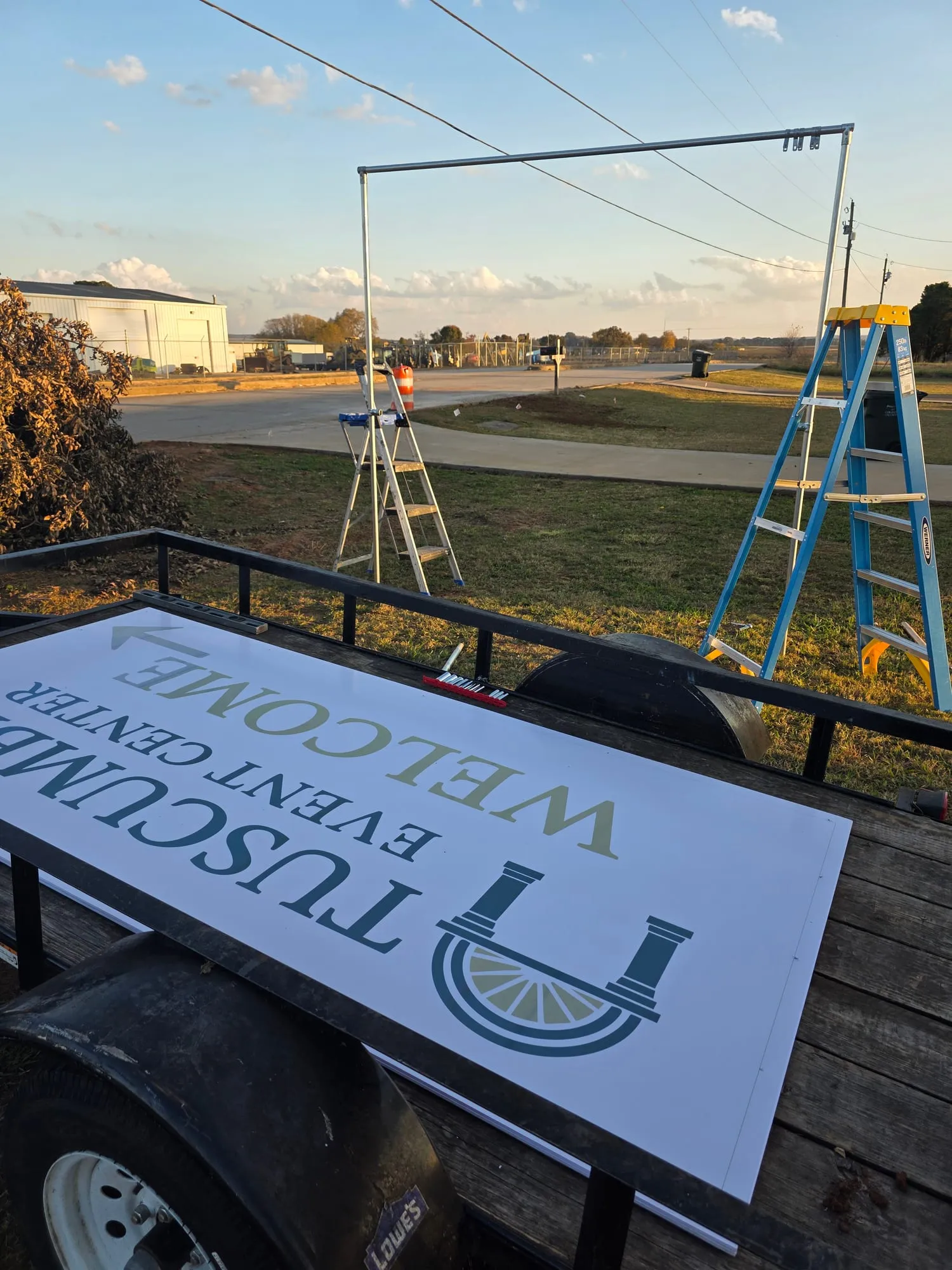 The printed banner panel lying on a trailer in front of the empty pipe frame, with two ladders standing in the grass.